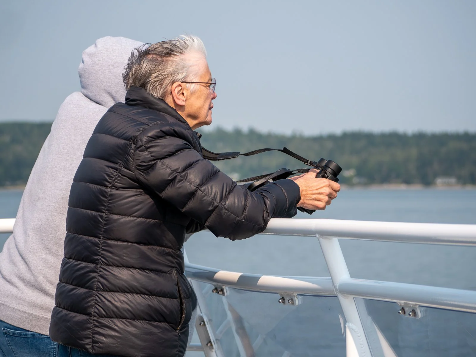 Older man with glasses in a black puffer jacket holding binoculars while on a boat, with a woman in a gray hooded sweatshirt standing next to him, overlooking a body of water with a distant shoreline.