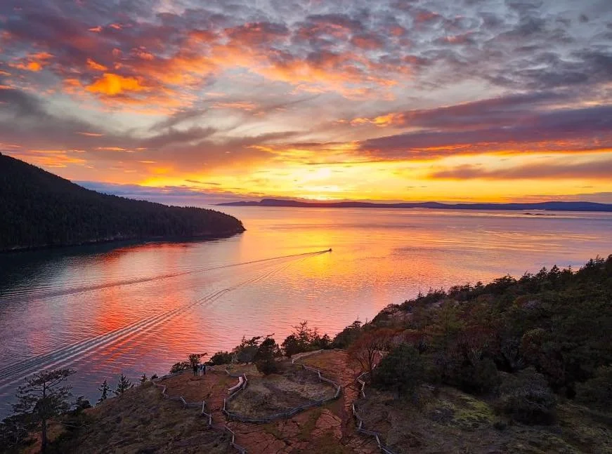 Sunset over a body of water with colorful clouds, a boat leaving a wake, and wooded shoreline and hills in the foreground.