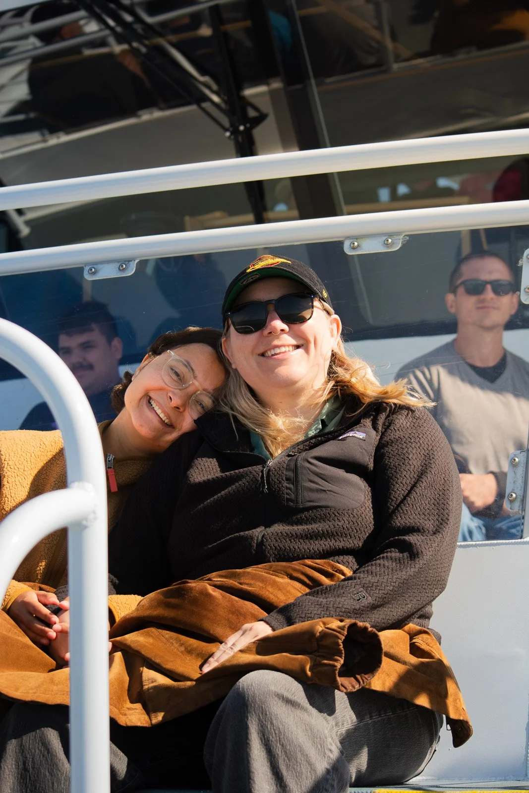 Two women smiling and sitting close together on an indoor train or tram, with a third man in the background wearing sunglasses, all enjoying a sunny day.