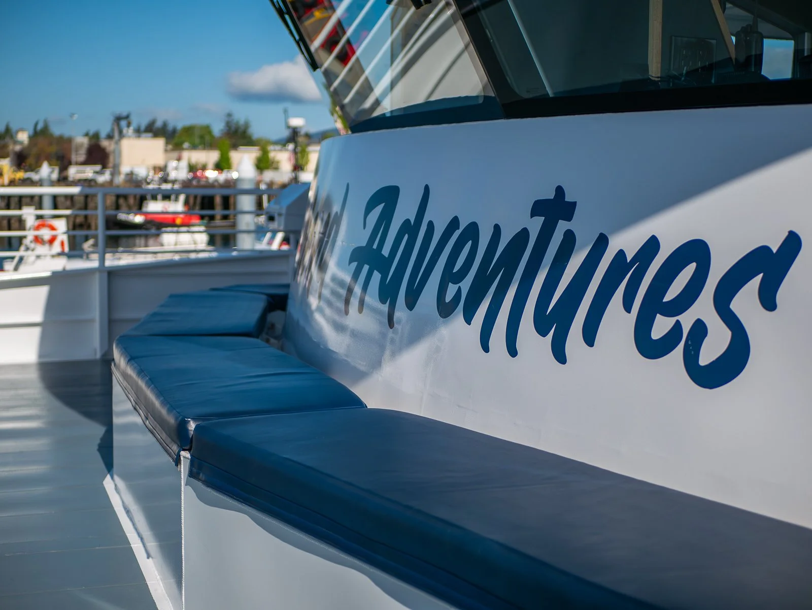 Close-up of a white boat with the name 'Adventurers' written in blue on the side, docked at a marina with other boats and a cityscape in the background, under a partly cloudy sky.