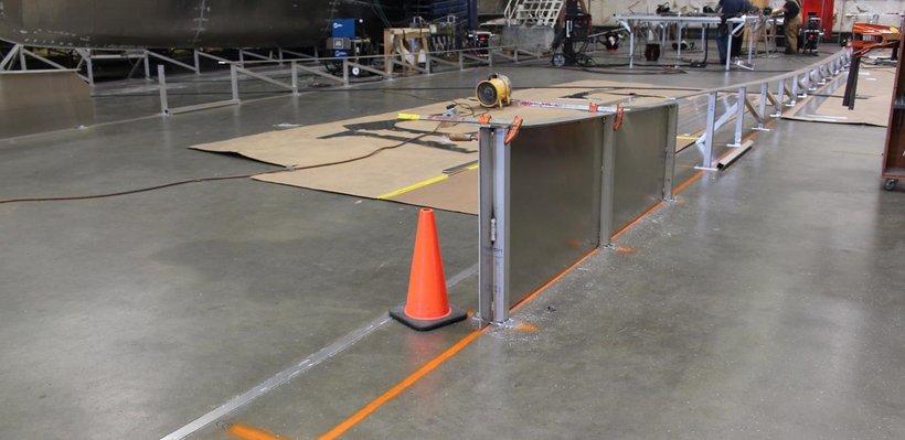 Indoor industrial workspace with construction or setup area, orange traffic cone, metal railing, and equipment on the floor.