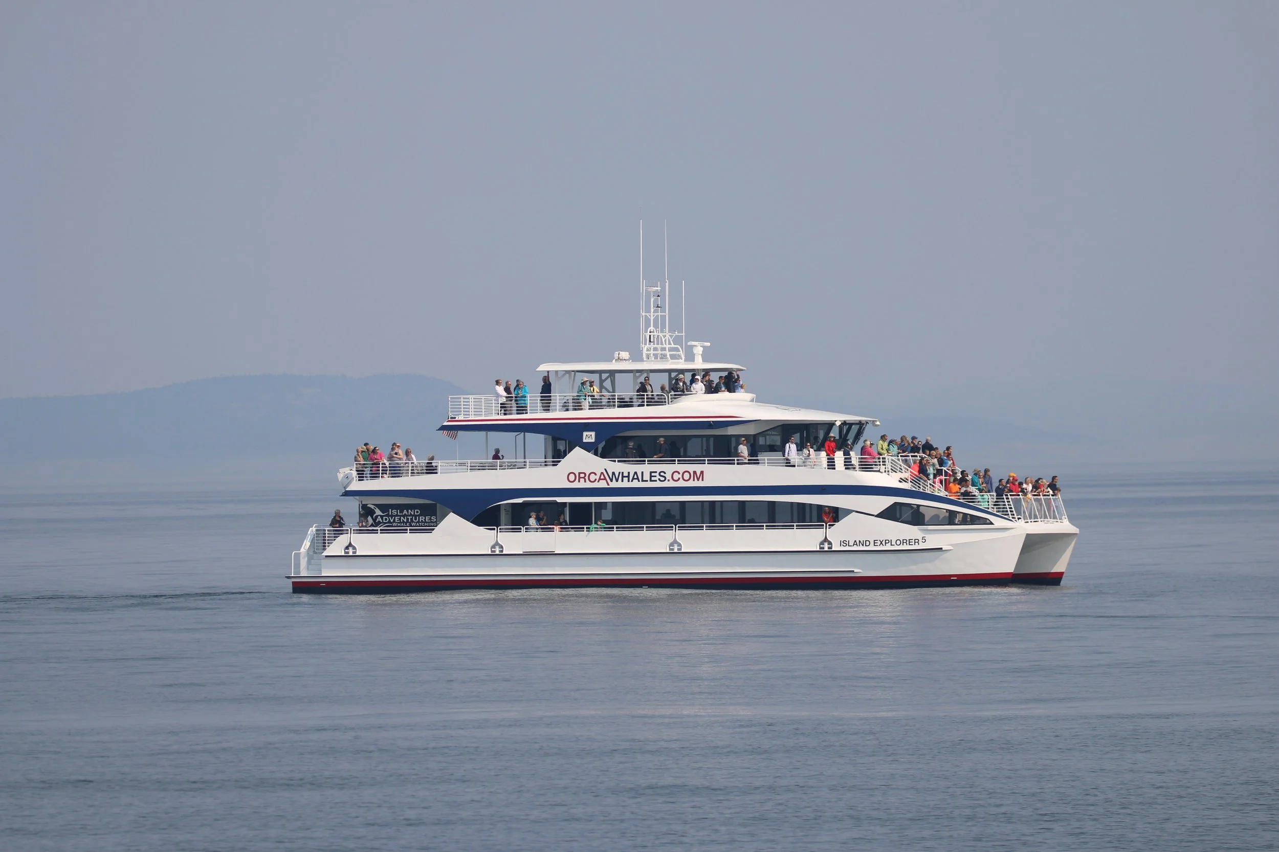A large white boat with multiple levels, carrying numerous passengers, is sailing on calm water under a grey sky. The boat has the words 'ORCA WHALES.COM' and 'ISLAND EXPLORER 5' written on its side.