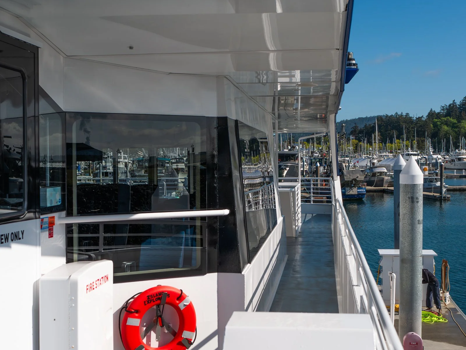 View of a marina with boats docked, taken from the deck of a boat, with clear blue sky.