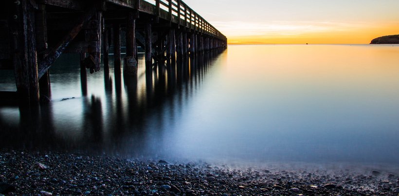 Sunset over calm water with a wooden pier extending into the distance, pebbled shoreline in the foreground.