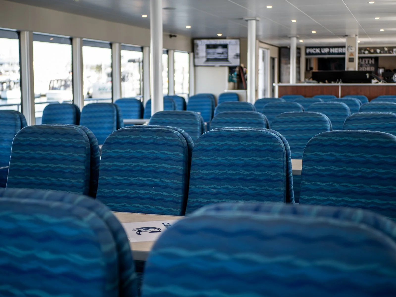 Empty blue patterned chairs inside a ferry or boat terminal with large windows and a view of boats outside.