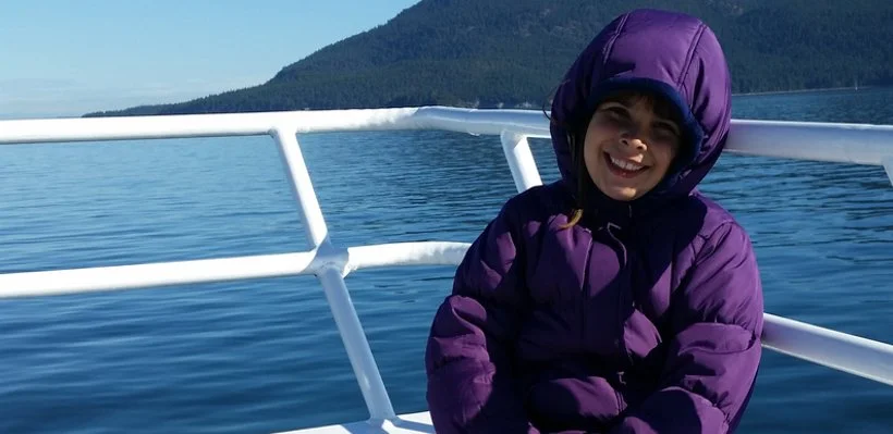 A young girl in a purple jacket with a hood, smiling on a boat with white railing, on a calm body of water with a mountain in the background.
