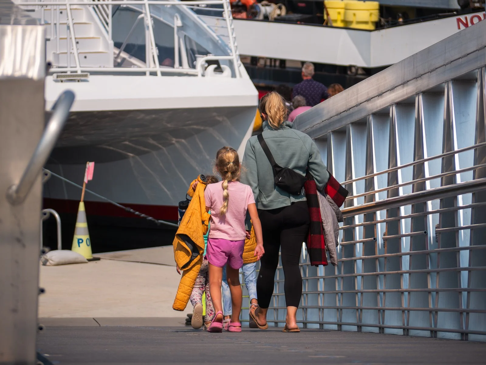 A woman and children walk on a dock towards a boat, with other passengers visible on the boat. The woman has a red and black plaid jacket and a black bag, and the children are carrying jackets and backpacks.