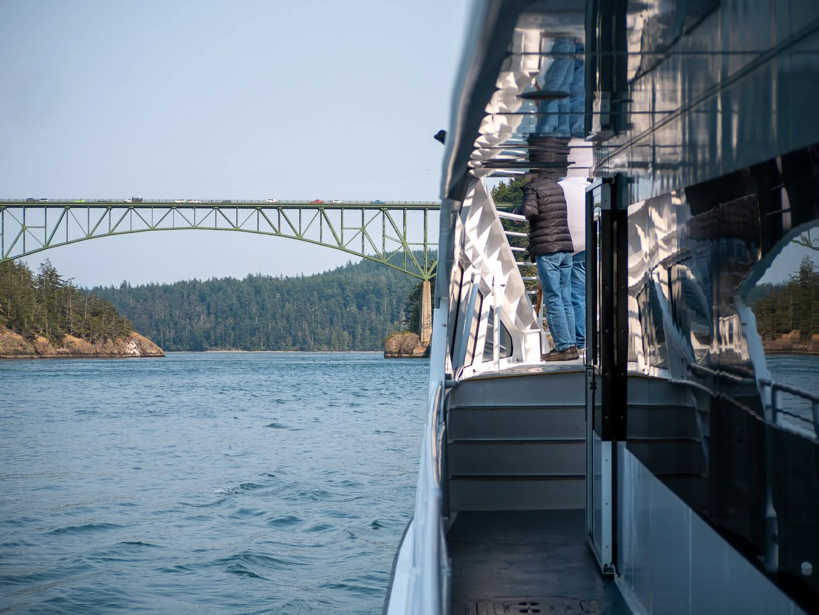 A view from a boat showing people on the deck looking at a bridge over water with a wooded shoreline in the background.