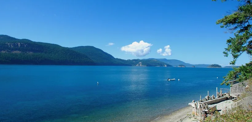 A serene lake with clear blue water, surrounded by mountains and green trees under a bright sky with a few clouds.