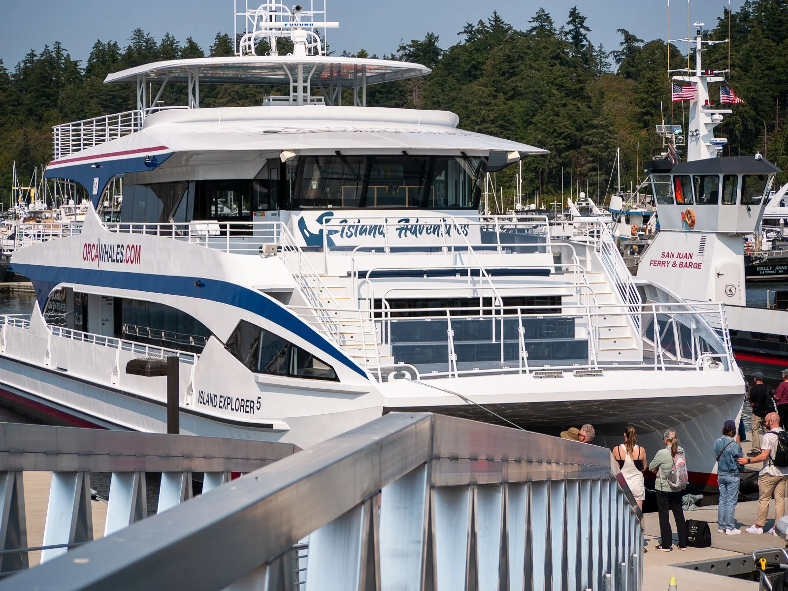 Passenger ferry boat docked at a port with people standing nearby and boats in the background, trees and a blue sky.