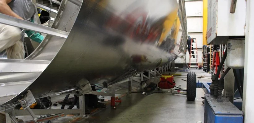 Inside a workshop or factory with machinery and tools, a large metallic cylindrical object is being worked on or assembled.