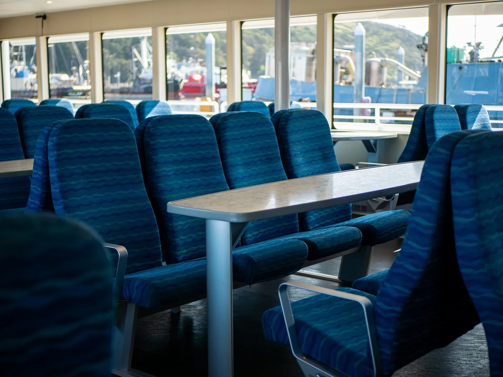 Empty passenger seats on a ferry with large windows, showing boats and water outside.