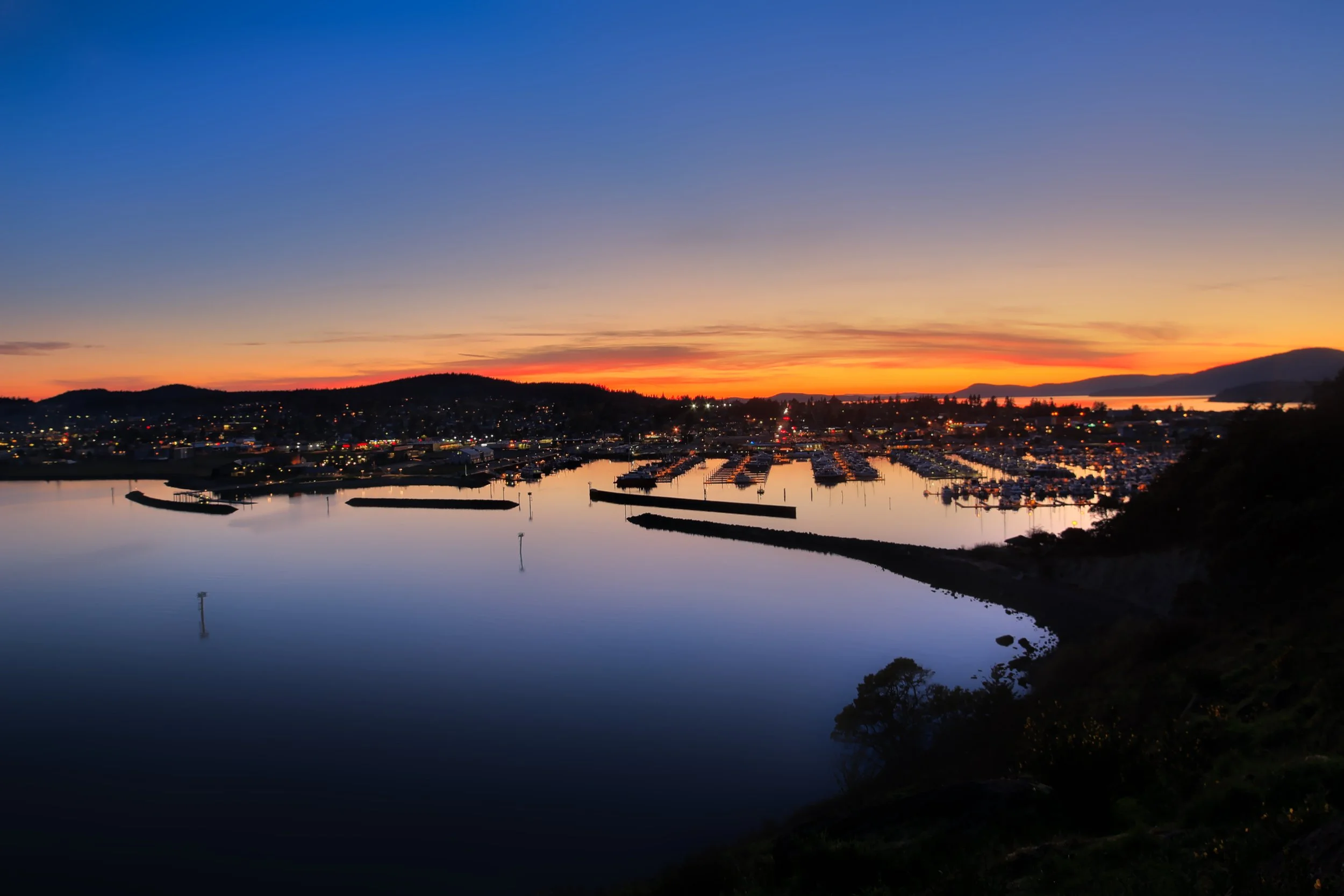 A scenic view of a waterfront town at sunset with colorful sky, calm water, marina with boats, and hills in the background.