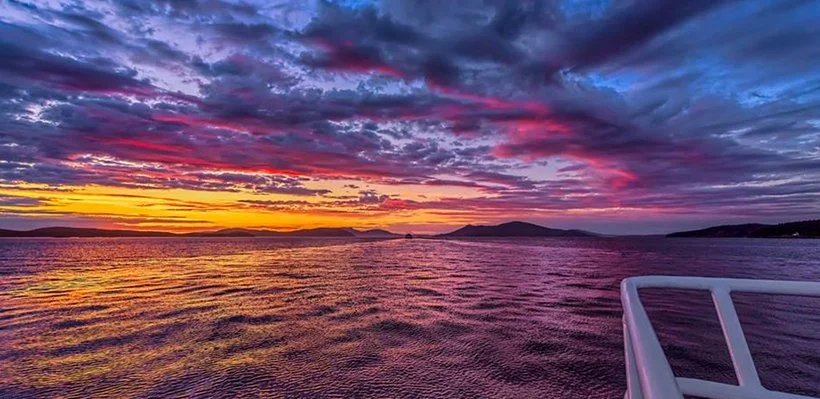 A vibrant sunset over a body of water with colorful clouds in the sky and silhouette of land in the distance, taken from a boat with part of the white railing visible.