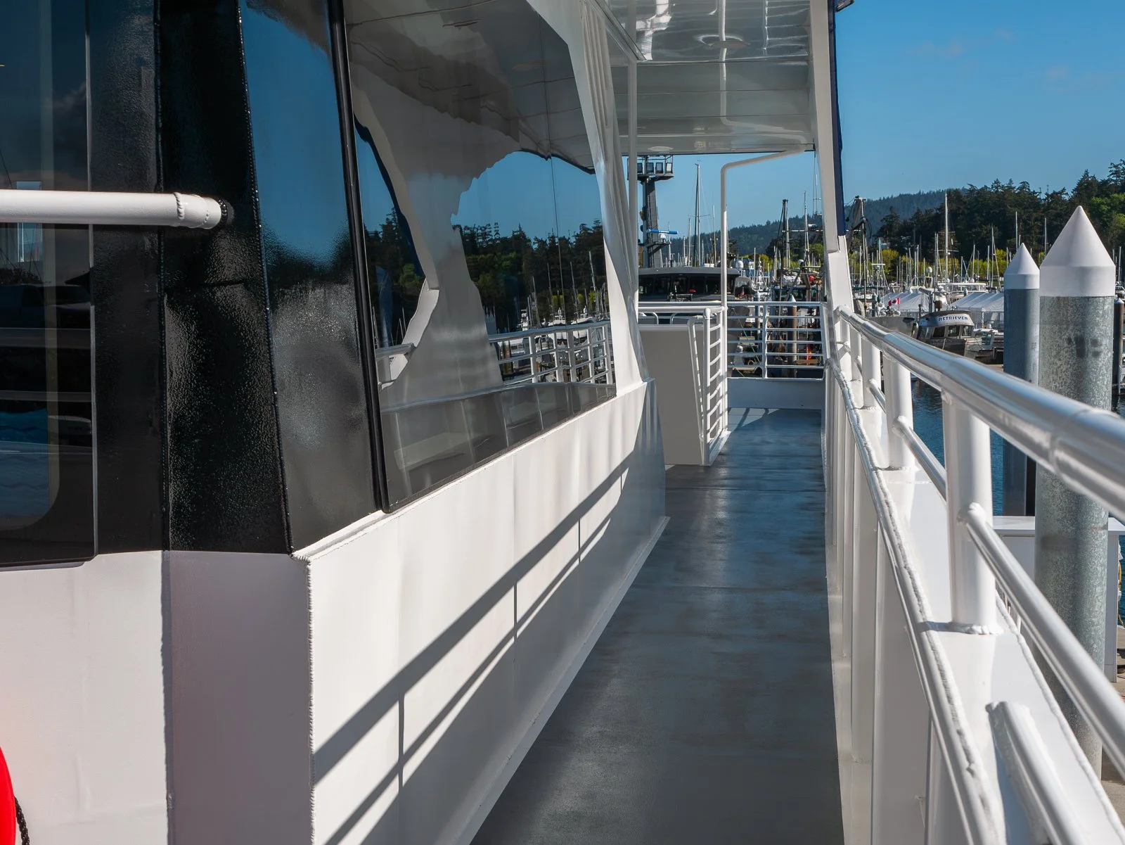 Side deck of a yacht with white railings, mainly white exterior with black accents, docked at a marina with multiple sailboats in the background under a clear blue sky.