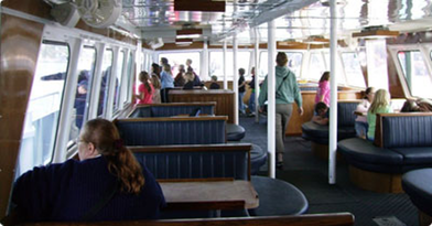 Interior of a ferry boat with passengers seated, windows on both sides, and a central aisle.
