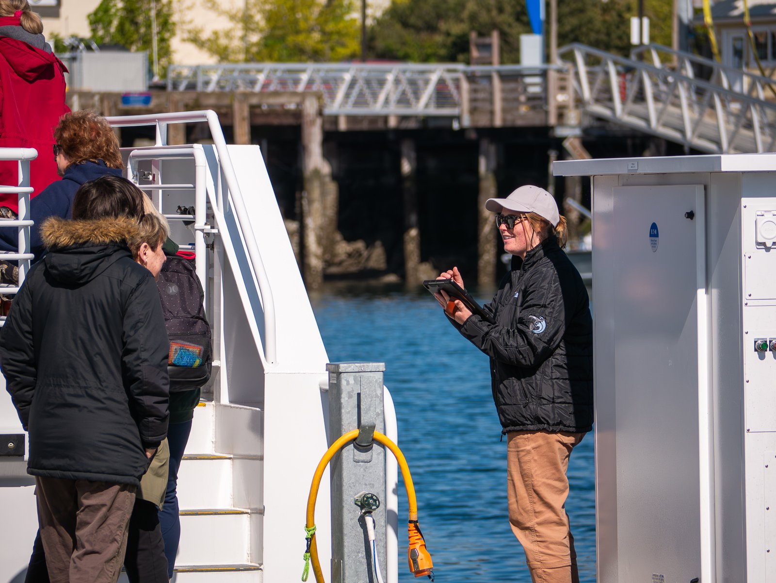 A woman in a black jacket, beige pants, and a white cap is talking to a group of people on a boat dock, near water, with a marina and wooden pier in the background.