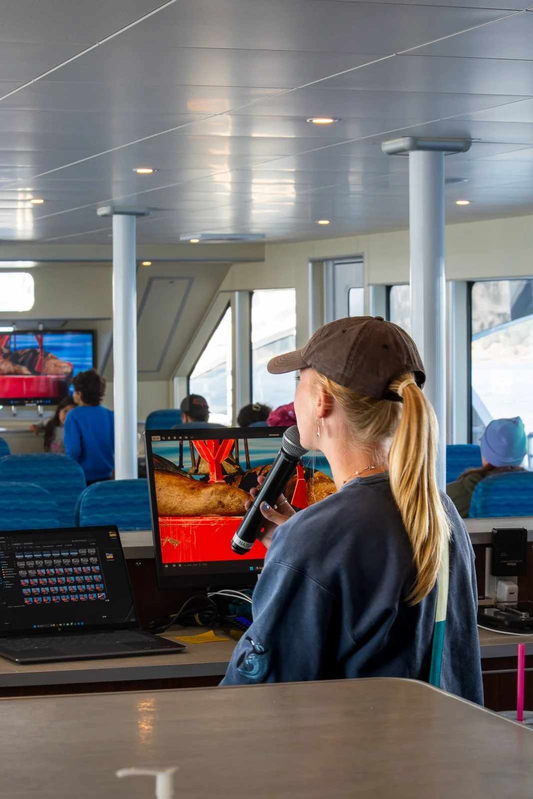 Woman with blonde hair, wearing a brown cap, speaking into a microphone, sitting in a boat or ferry's control room or passenger area, with a large window showing an outdoor scene and other passengers in the background.