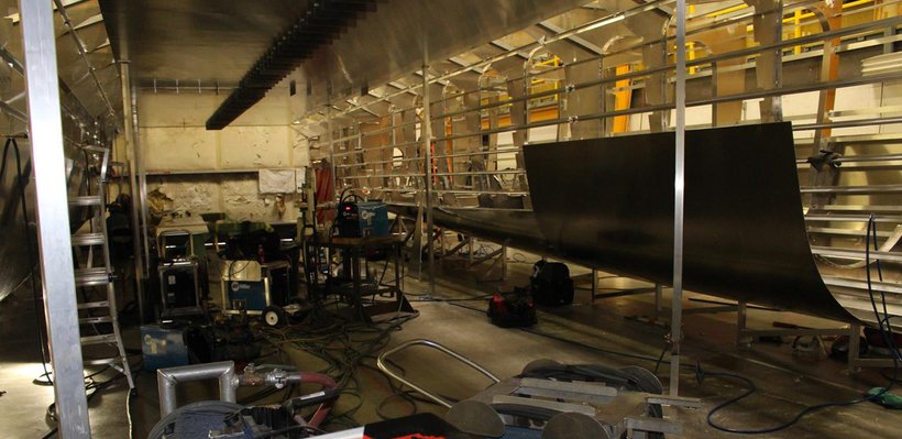 Industrial workspace with metal framework, a large partially assembled structure, and various tools and equipment scattered around.