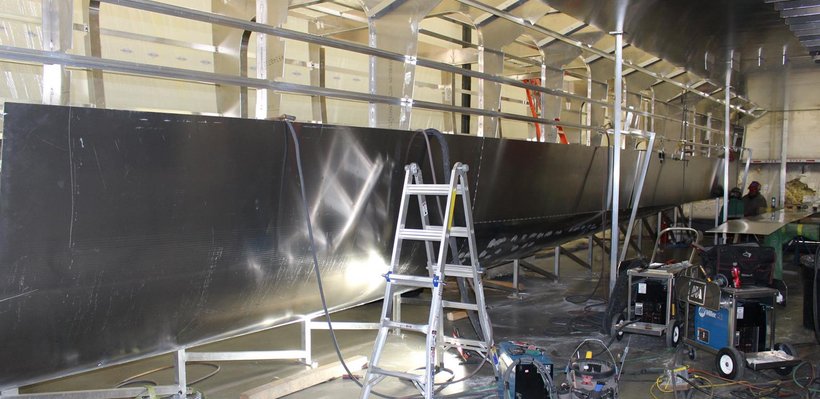 Large boat under construction inside a workshop, with scaffolding, ladder, and workshop tools nearby.