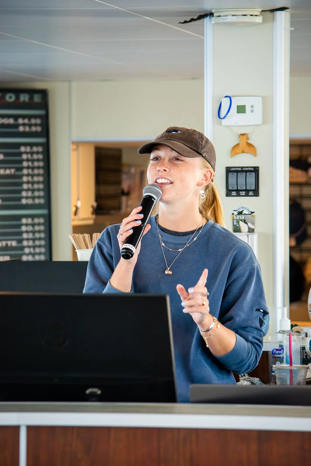 Young woman wearing a brown cap and blue sweatshirt speaking into a microphone at a shop counter.