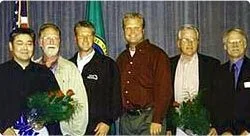 Group of six men standing together indoors, some holding bouquets of flowers.