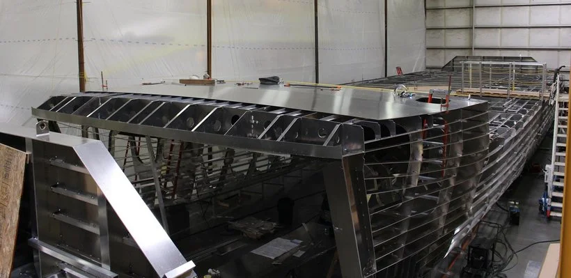 A large metallic boat under construction inside a workshop, with visible ribs and framework in progress.