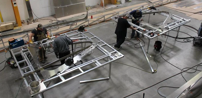 Four workers assembling large metal frames or structures on a workshop floor surrounded by tools and equipment.
