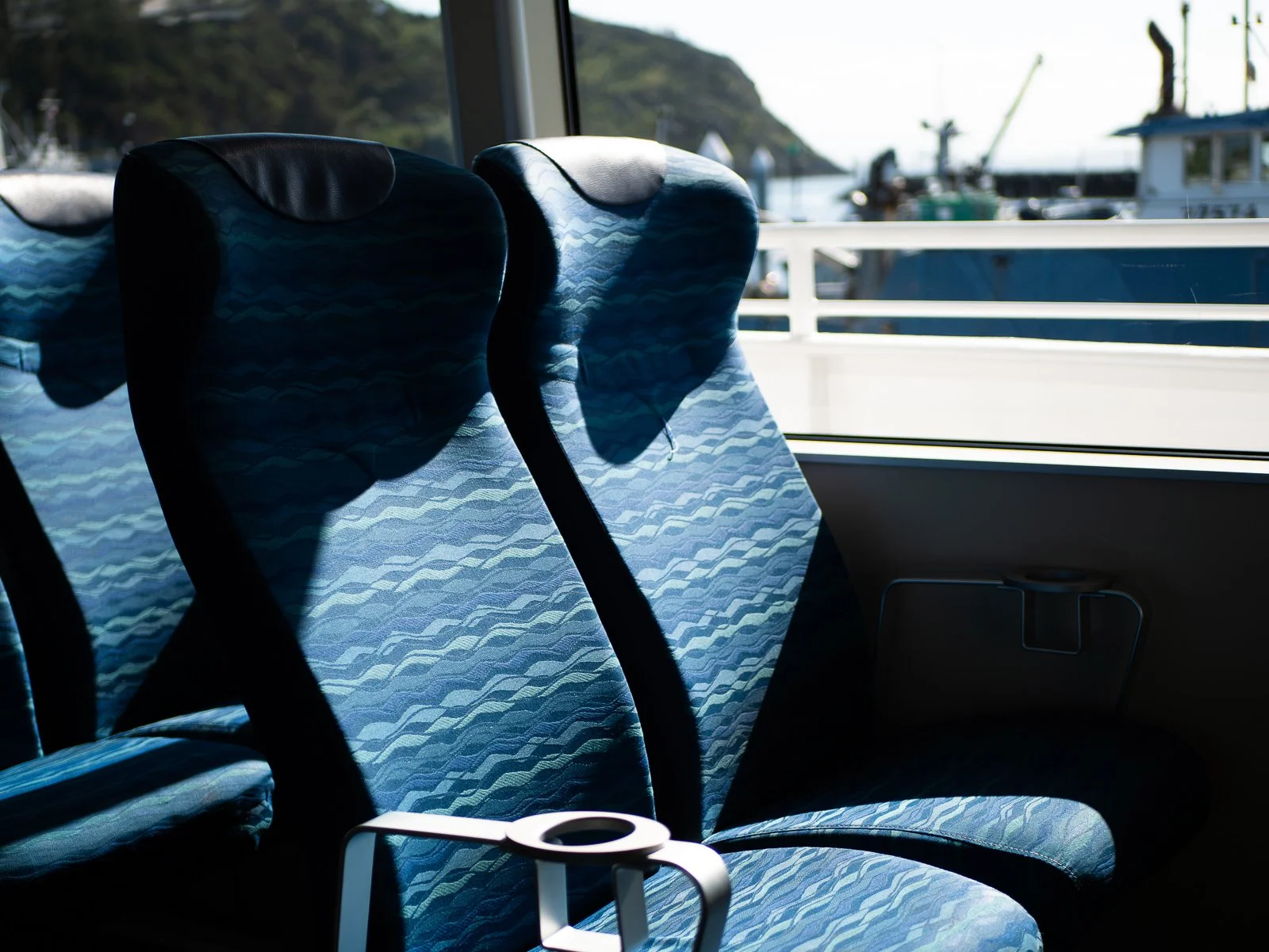 Empty blue patterned bus seats next to a window showing a marina with boats and hills.