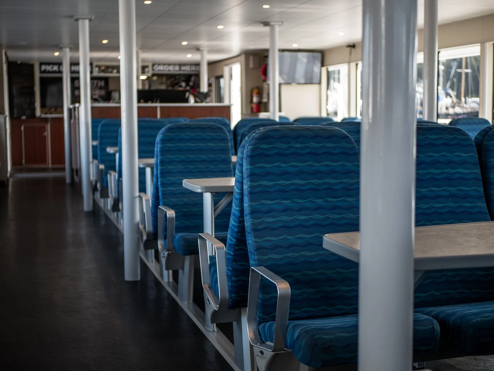 Interior of a ferry with empty blue patterned seats, tables, large windows, and a flat-screen TV, with boats visible outside.