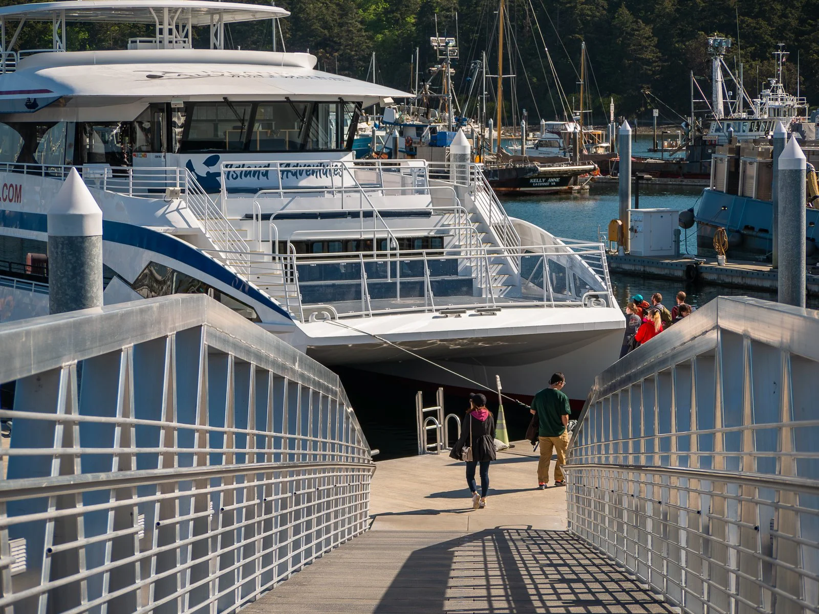 People walking down a ramp onto a large white boat docked at a marina, with other boats and sailboats in the background.
