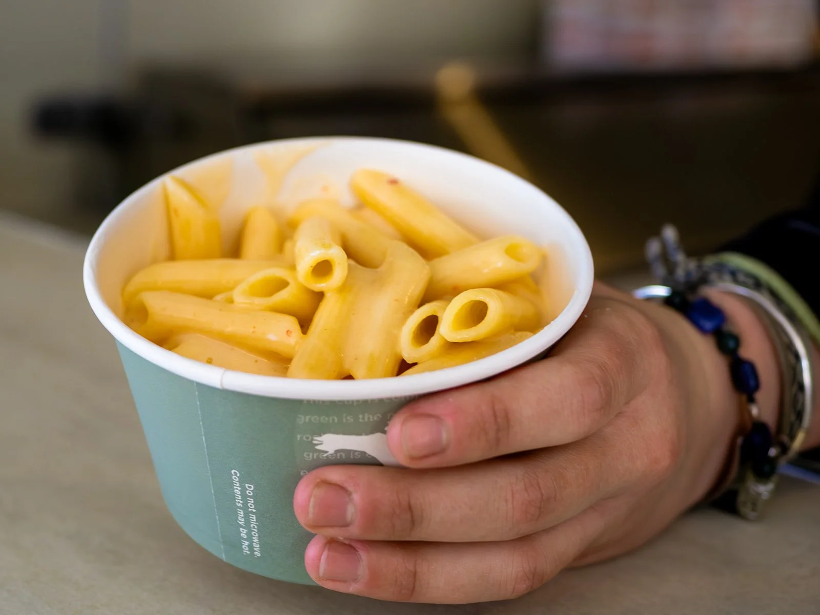 A hand holding a paper cup filled with macaroni and cheese.
