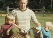 A man with two young girls outdoors on a sunny day, sitting on grass near a wooden fence.