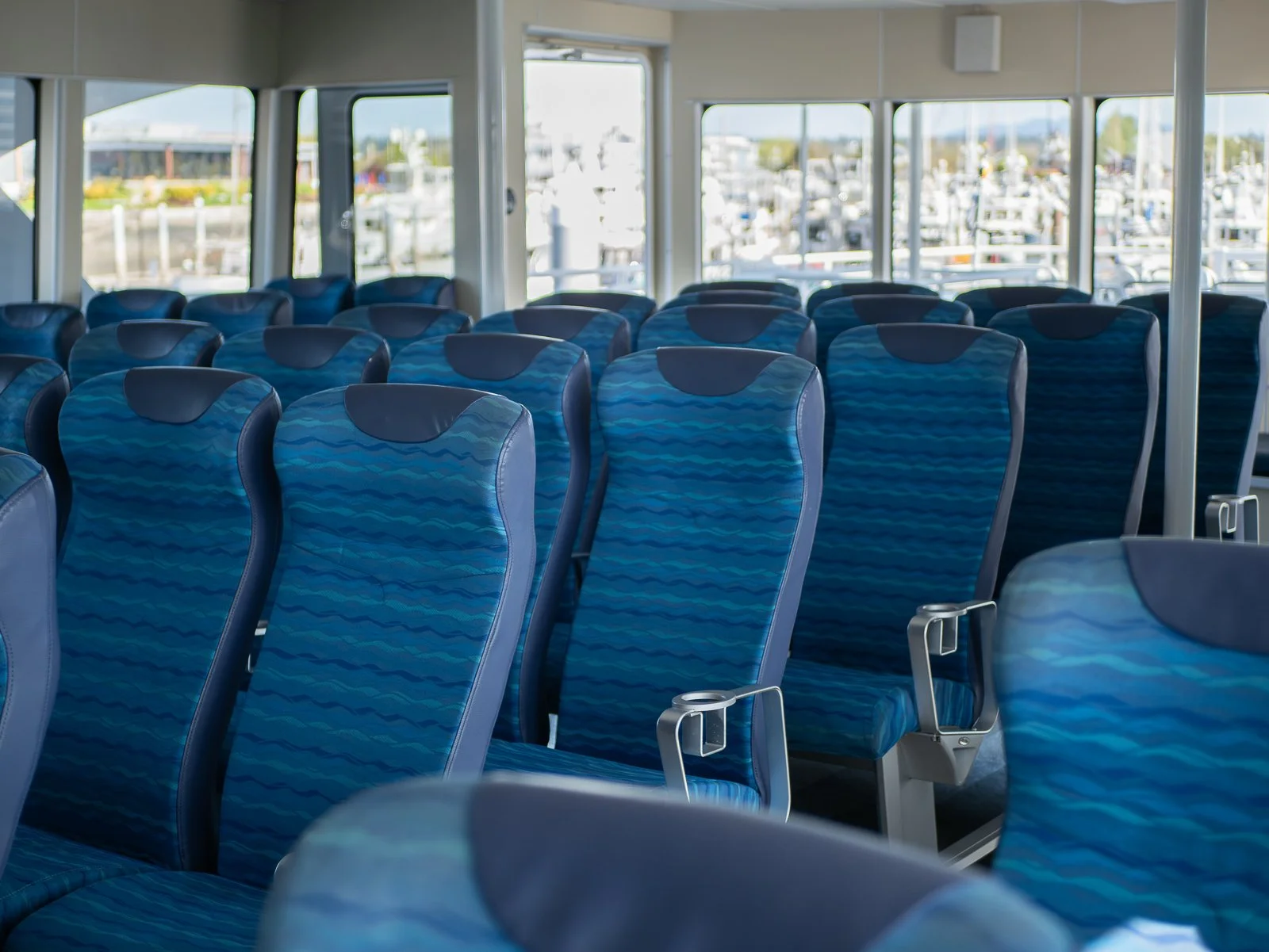 Empty blue padded seats inside a boat's passenger area, with large windows showing a marina with boats docked outside.