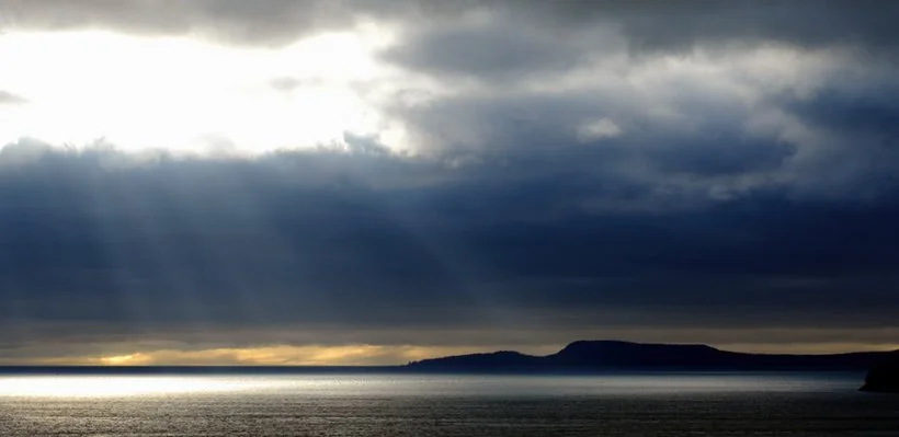 Dark storm clouds over the ocean with rays of sunlight breaking through, and a landmass in the distance.