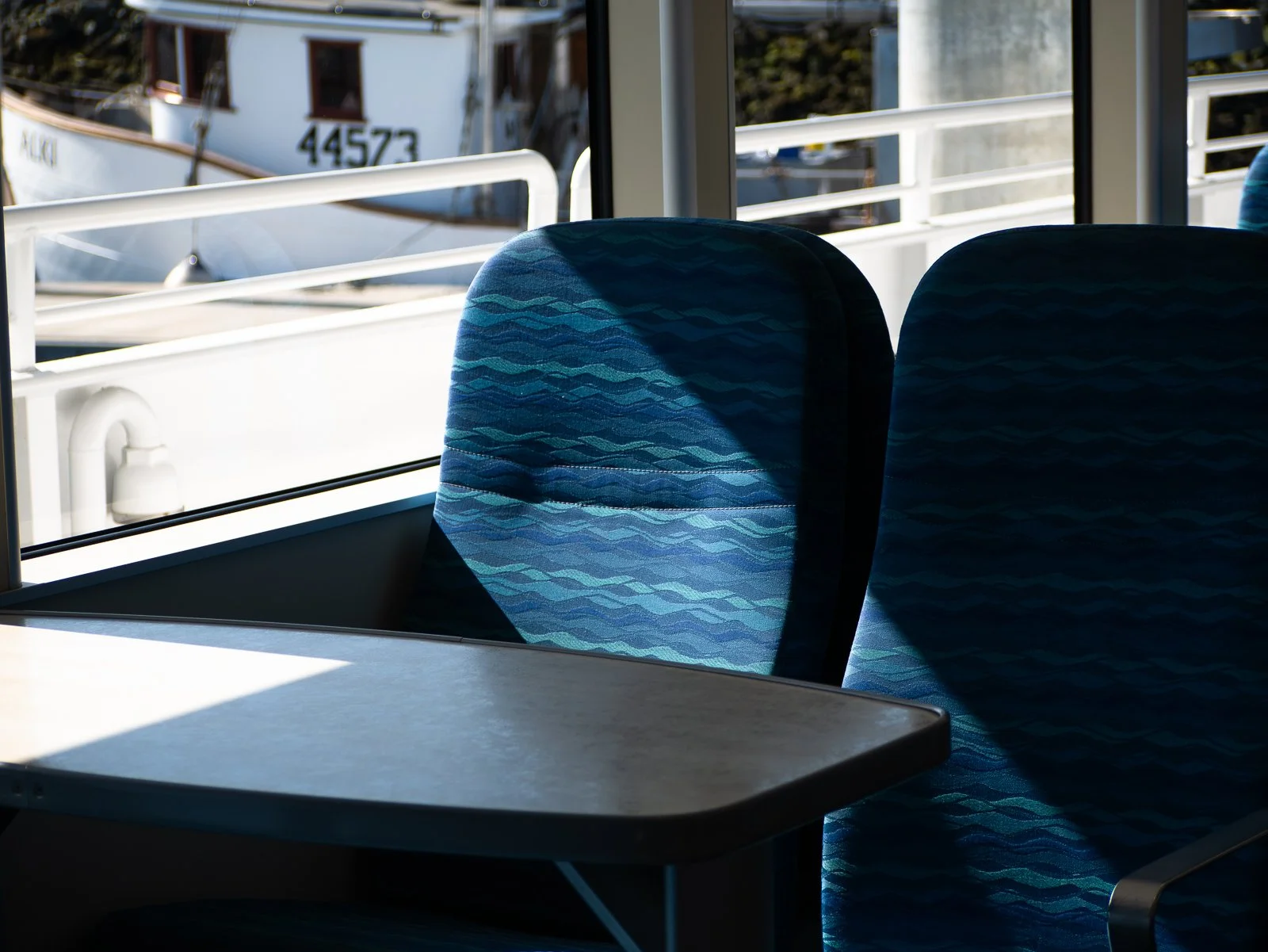 Empty blue patterned seat and table inside a ferry or bus, with sunlight casting shadows, and boats outside through the window.