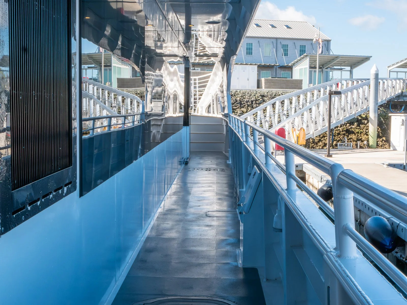 View of the side of a modern industrial or transportation vessel with a walkway and safety railings, possibly part of a ferry or cruise ship, with buildings and sky in the background.