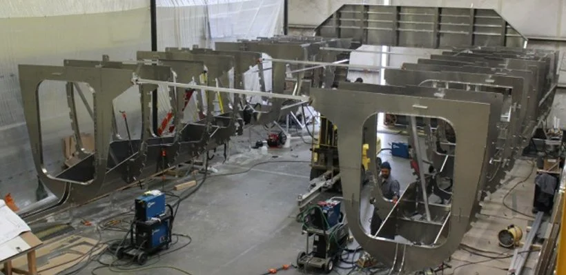 Large metal frames, possibly aircraft sections, are being constructed or assembled in a factory or industrial setting with workers and tools around.