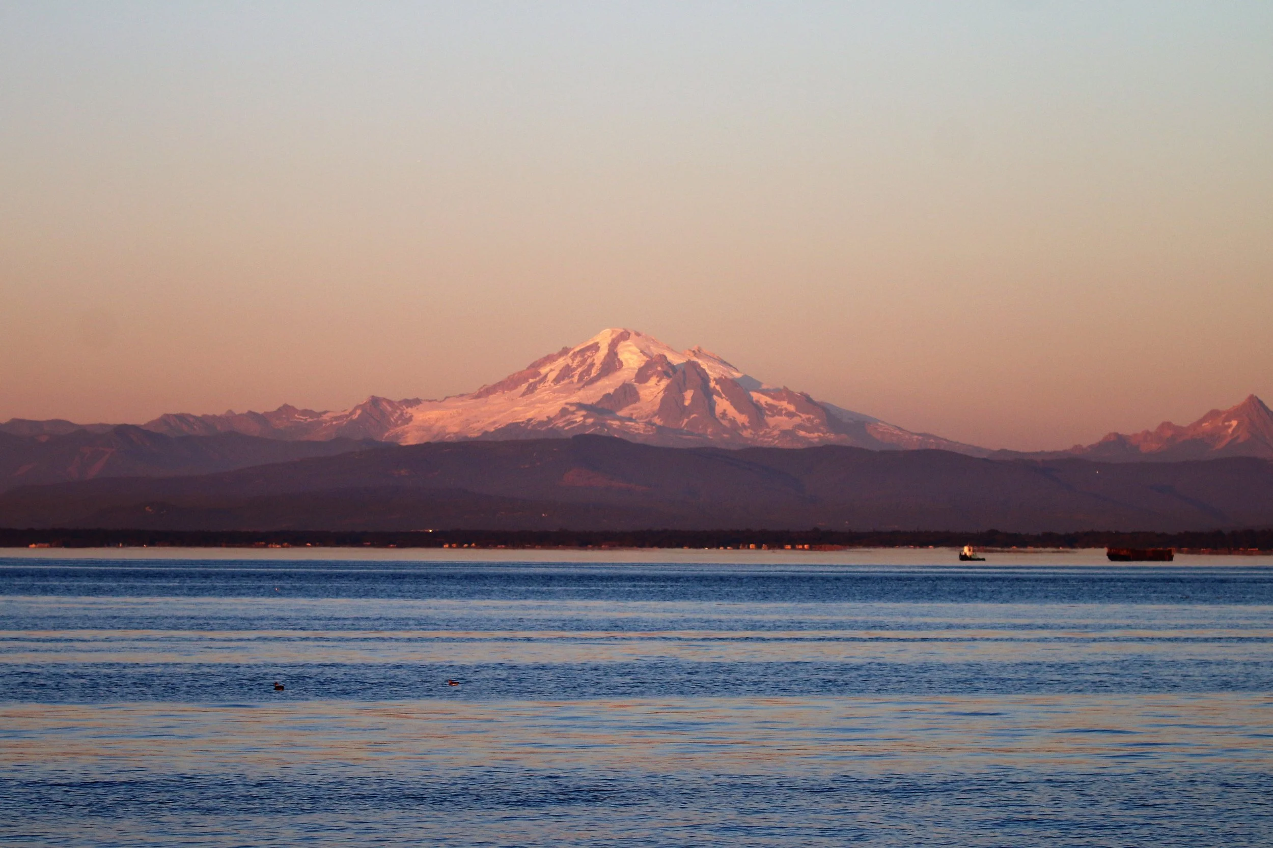 Snow-capped Mount Rainier viewed across a body of water with a small boat in the foreground during sunset.