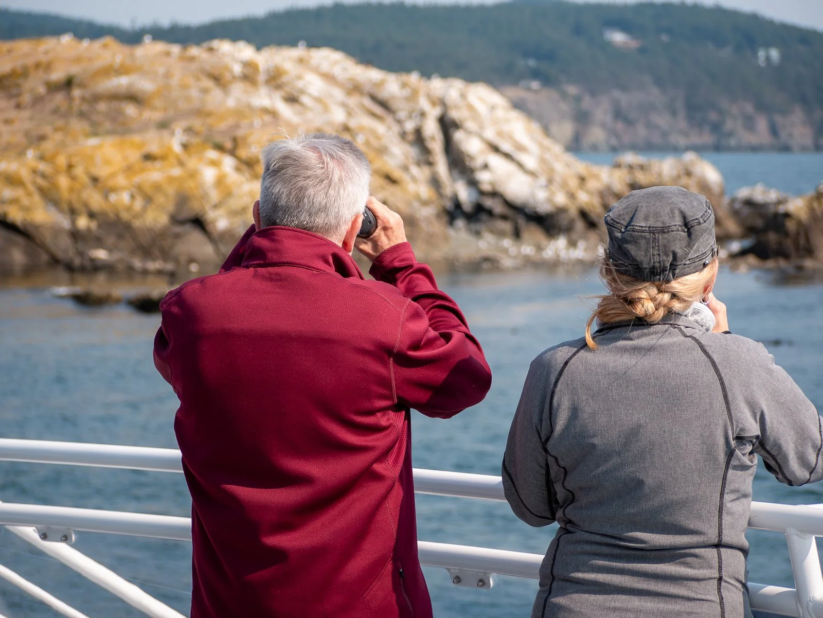Two people standing on a boat looking at ocean scenery. One man with gray hair in a red jacket is using binoculars, while a woman with blonde hair in a gray cap and jacket is taking a picture.