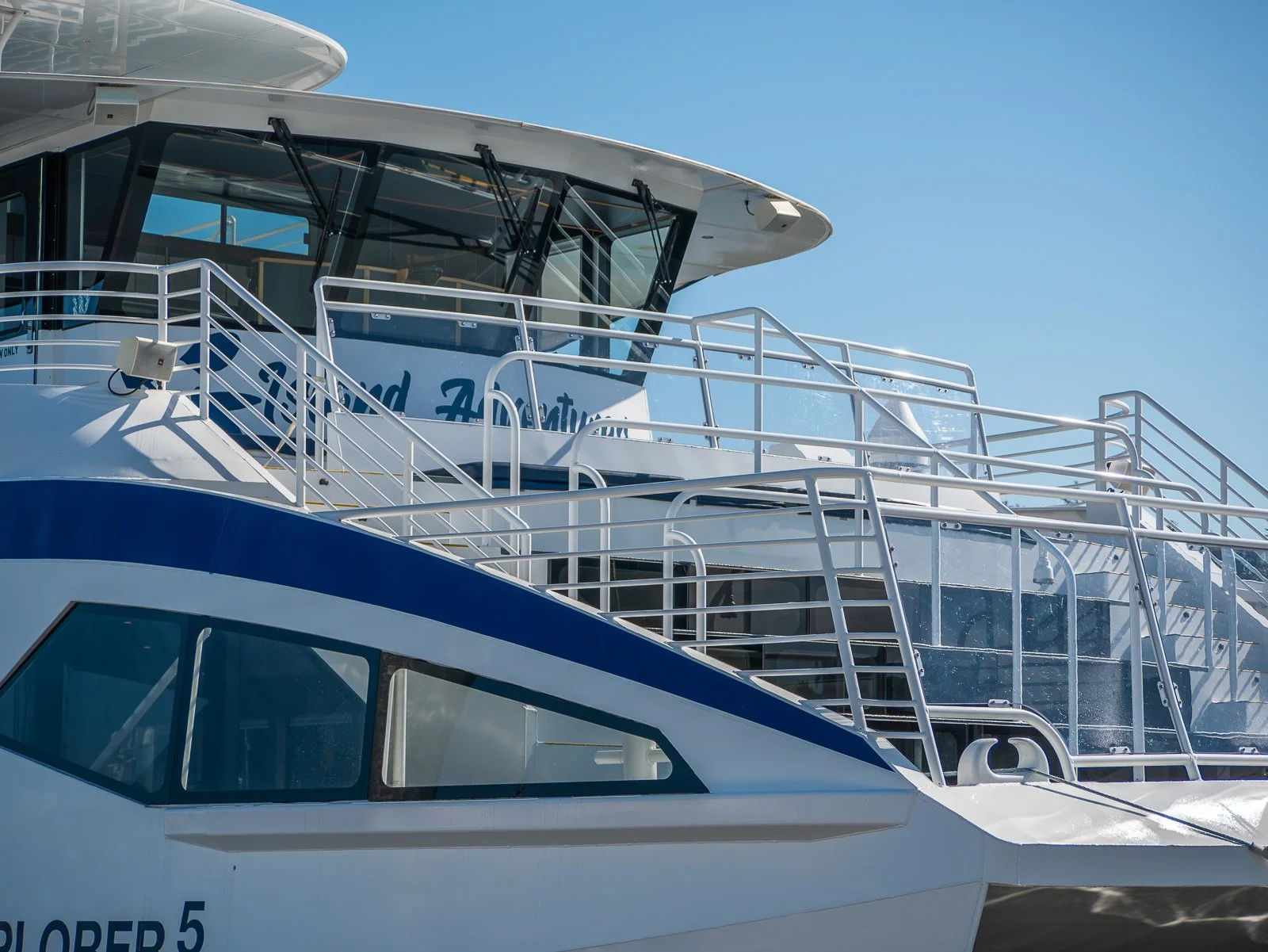 Close-up view of the upper deck of a white and blue yacht with multiple railings, windows, and a partially visible sign reading 'and Fa...' under a clear blue sky.