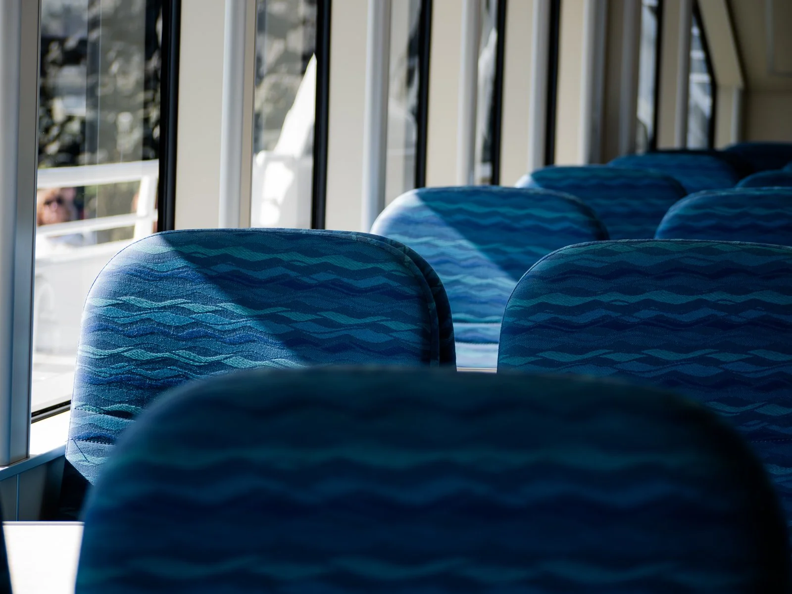Empty blue patterned bus seats beside large windows with sunlight and a blurred exterior view.