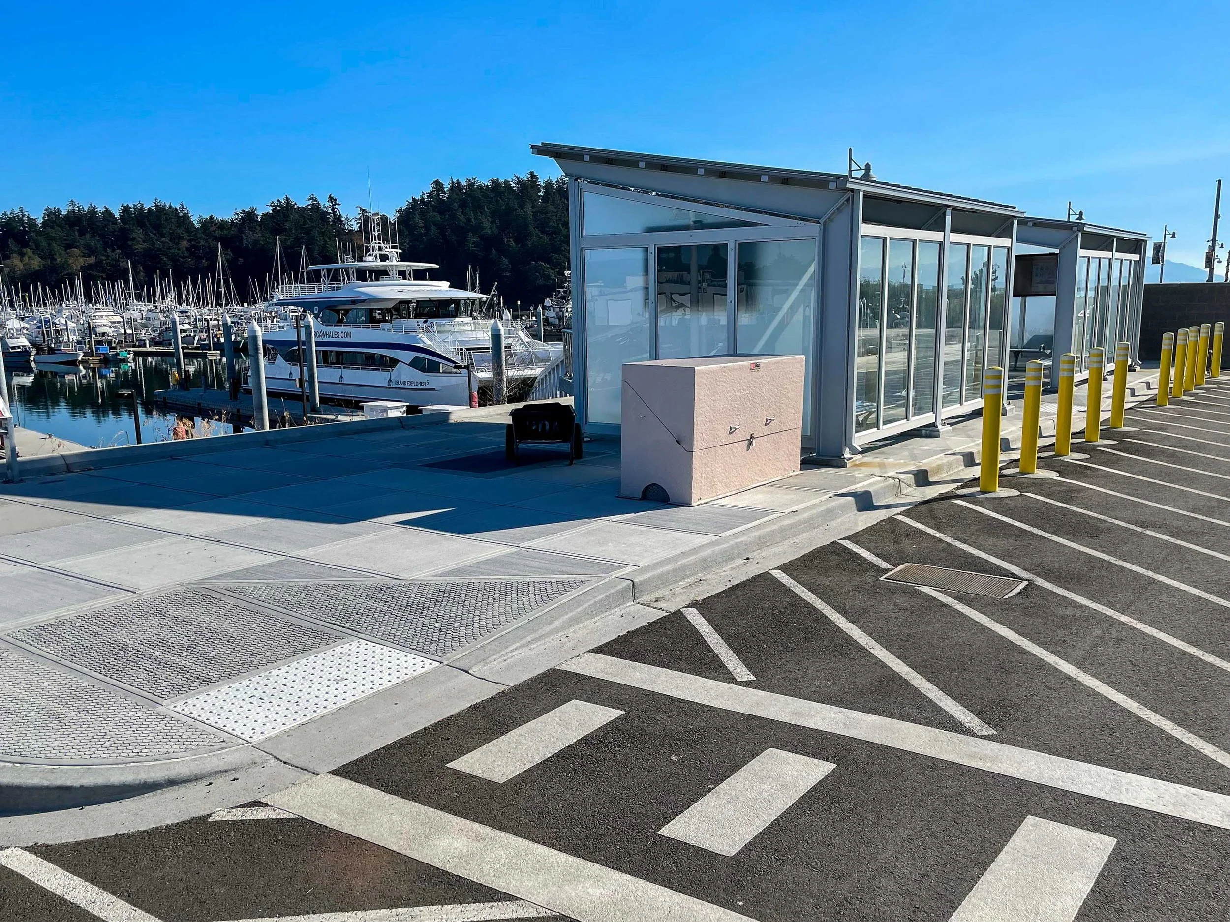 Marina with boats docked, modern glass shelter, parking lot with painted lines and yellow bollards, forested hillside in the background under a clear blue sky.