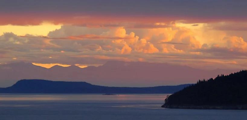 A scenic view of a large body of water with forested islands and distant mountains under a colorful sky with clouds at sunset.