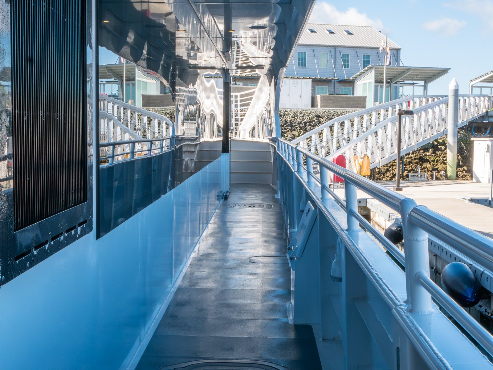 View from the deck of a boat showing a blue walkway with metal railings, leading to the interior, with buildings and sky in the background.