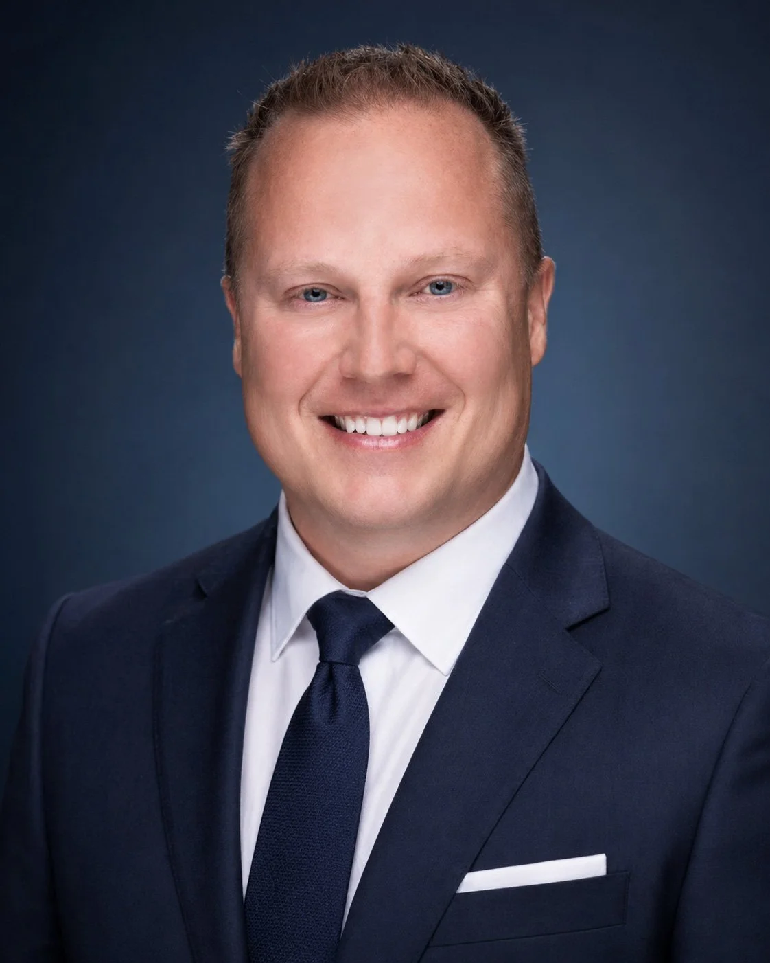 Headshot of a smiling man in a navy suit, white shirt, and navy tie with a dark blue background.