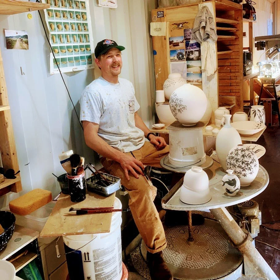 A man sitting at a pottery wheel in a ceramics studio, surrounded by white ceramic vases and bowls, with shelves and tools in the background.