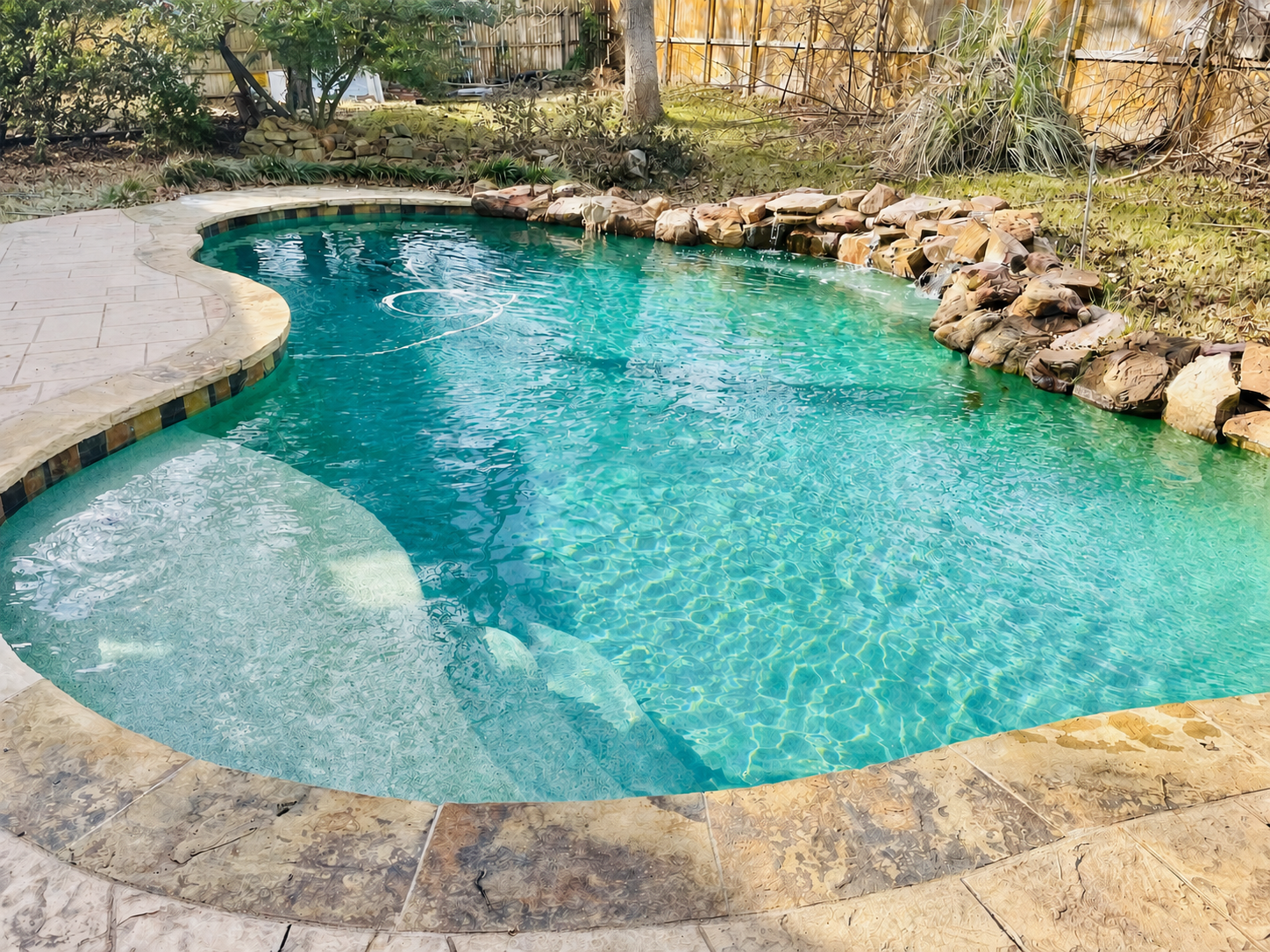 A backyard swimming pool with clear blue water, surrounded by stone and tile decking, rocks, and plants along the fence line.