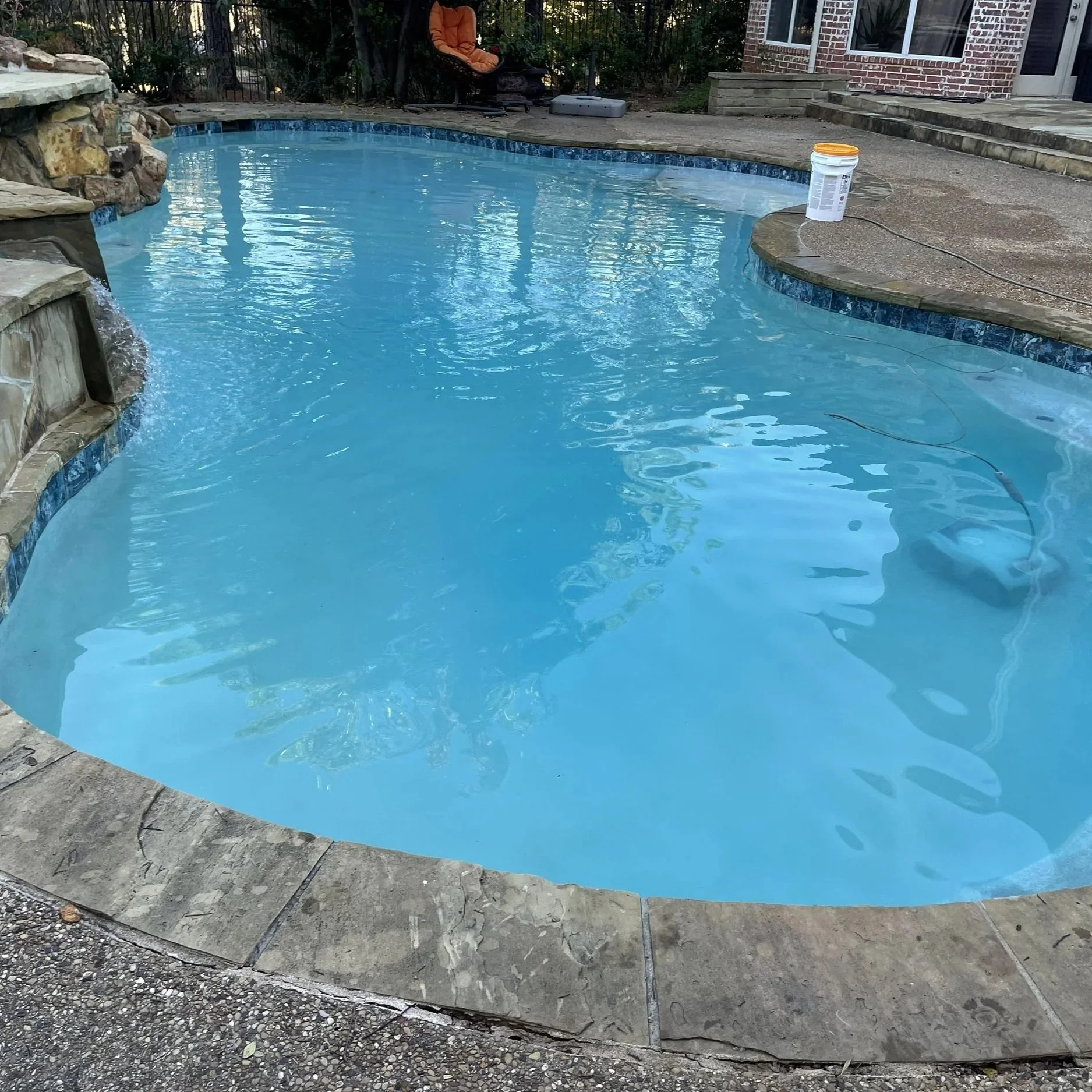 A backyard in-ground swimming pool with blue water, surrounded by stone and concrete decking, with a bucket, a chair, and a house in the background.