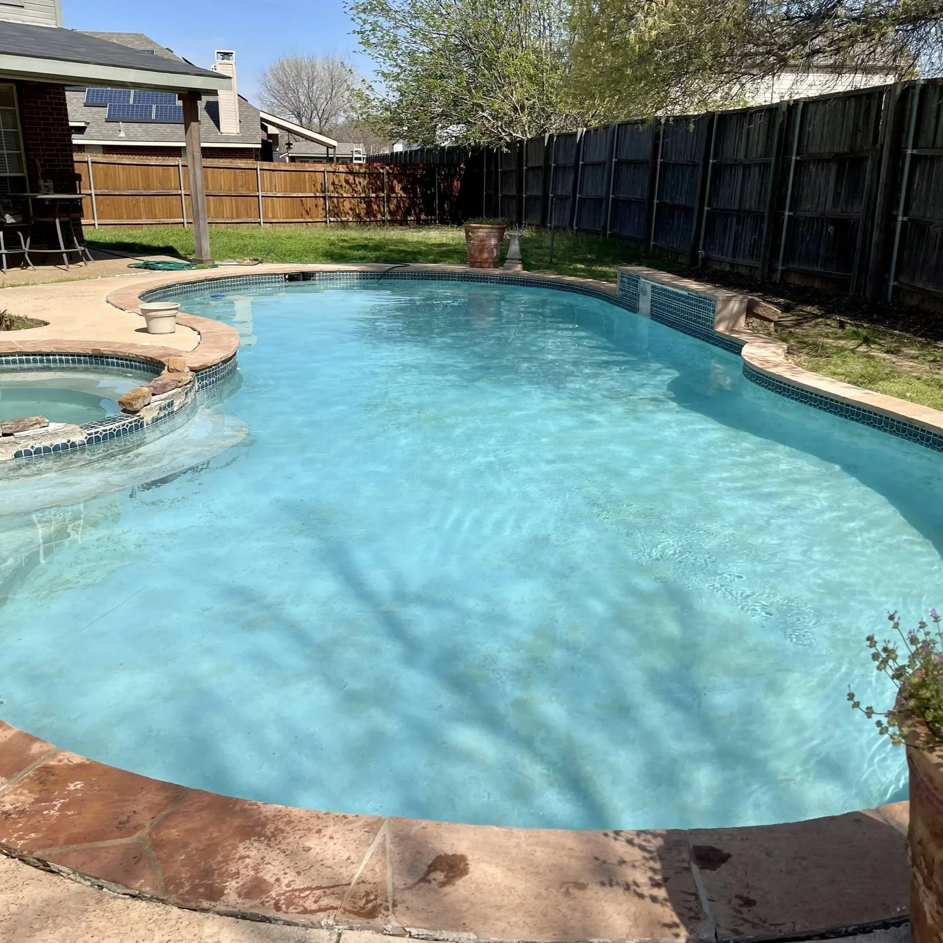 A backyard with a swimming pool and a hot tub, surrounded by a brick patio. There is a wooden fence and a house with solar panels on the roof. Trees and shrubs are visible in the background.